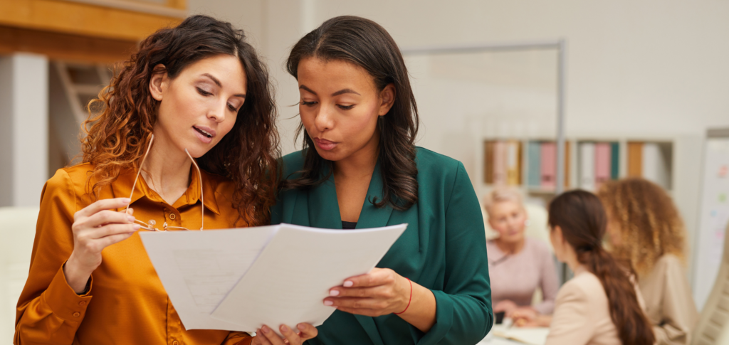 Two Women Looking at Papers and Discussing Business While Colleagues Work at a Table Behind Them