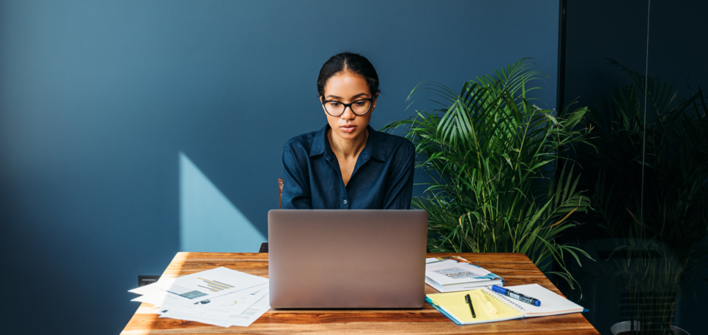 Young Professional Woman Concentrating and Working on a Laptop Surrounded by Documents, Notebooks, and Pens